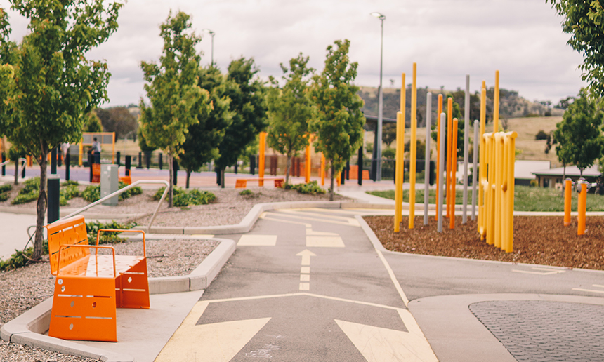 Playground and park with colourful bench seats, and play equipment with green trees and directional signage painted on paths.